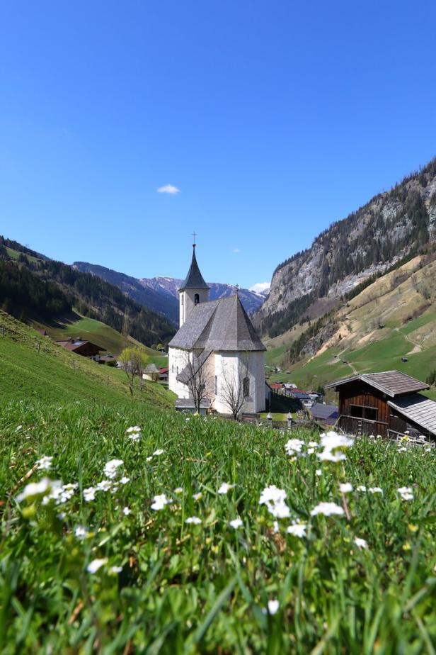 Eine Kirche steht in einer grünen Wiese vor einer Bergkulisse.