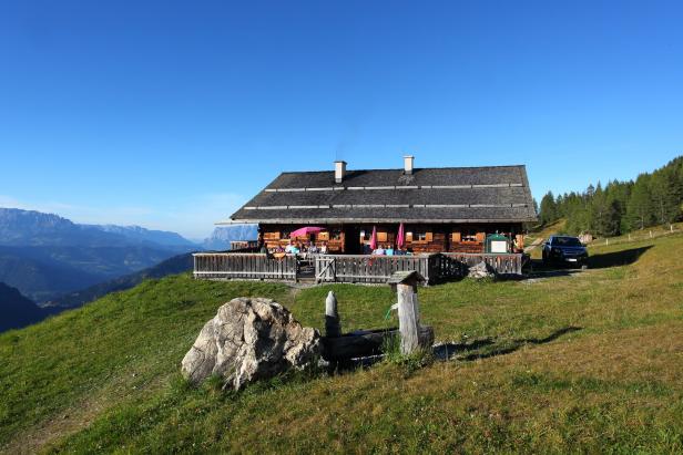Ein hölzernes Berghaus mit Gästen auf der Terrasse vor einer Bergkulisse.