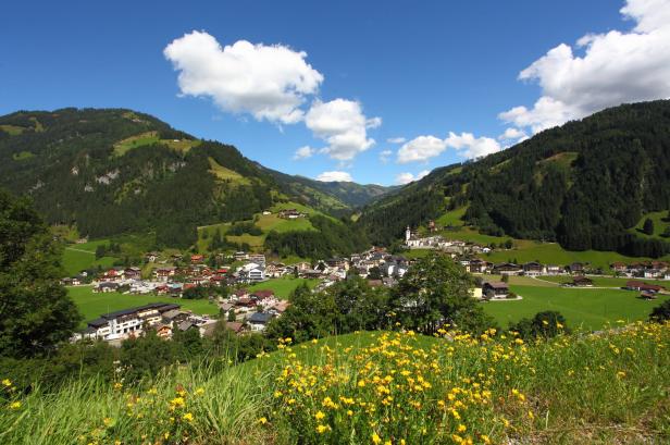 Ein Dorf liegt eingebettet in eine grüne Hügellandschaft unter blauem Himmel.