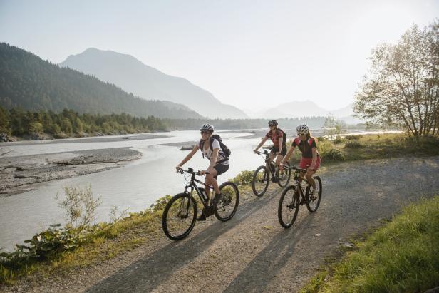 Drei Frauen fahren mit Mountainbikes auf einem Weg entlang eines Flusses vor einer Bergkulisse.