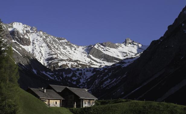Holzhütten vor einer Bergkulisse mit schneebedeckten Gipfeln.