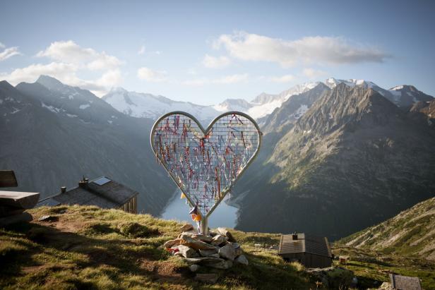 Ein herzförmiges Gitter mit bunten Bändern vor einer Berglandschaft.