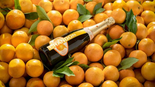Eine Sektflasche mit einer Orange auf dem Etikett liegt auf einem Haufen Orangen.