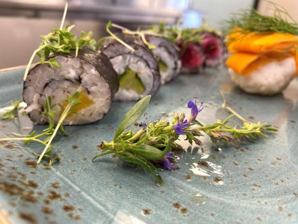 Gemüsesushi auf einem hellblauen Keramikteller angerichtet mit verschiedenen Kräutern und Blüten.