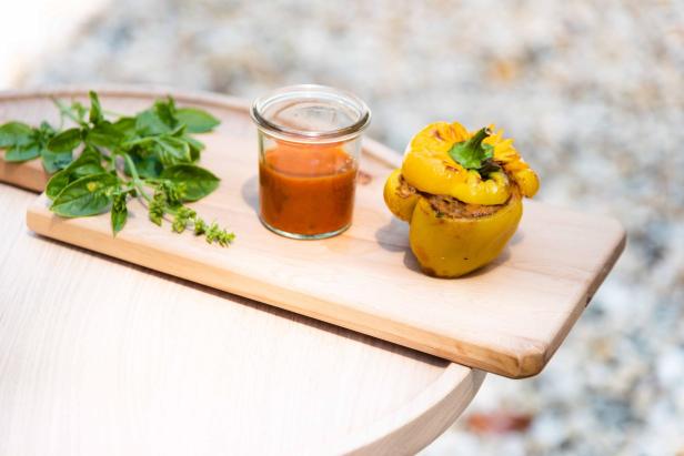 In einem kleinen runden Weck-Glas ist Gazpacho, daneben befindet sich eine gelbe, gefüllte Paprika und Kräuter auf einem Holzbrett.