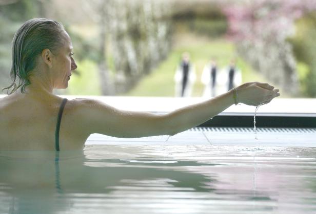 Eine Frau mittleren Alters ist in einem Pool, sie lässt das Wasser durch ihre Finger rinnen, während sie den Arm hebt.