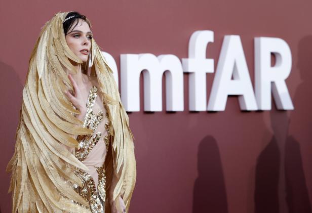 Eine Frau in einem Kleid mit goldenen Federn posiert vor dem Hintergrund des „amfAR“-Logos.