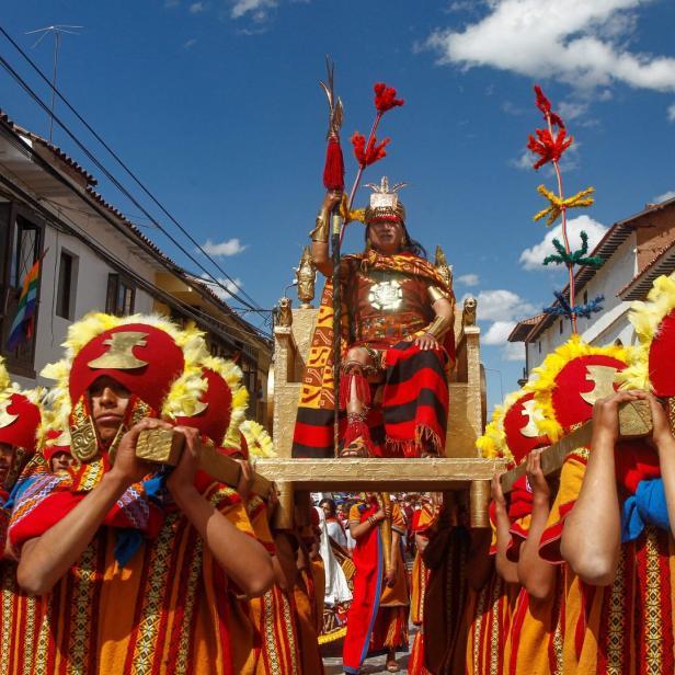 Eine Gruppe von Menschen trägt einen Mann in traditioneller Inka-Kleidung auf einem goldenen Thron durch eine Stadt.