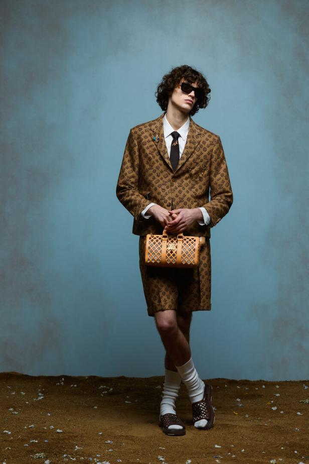 Ein Mann mit Sonnenbrille trägt einen braunen Anzug mit Shorts und hält eine Handtasche.
