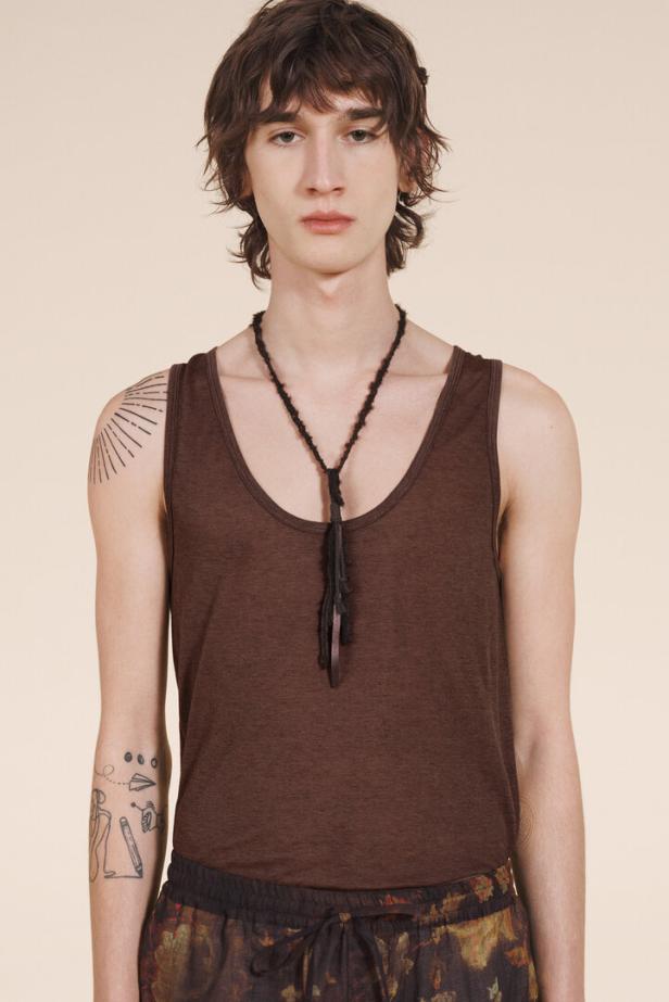 Ein Mann mit braunem Tanktop und Halskette posiert vor einem hellen Hintergrund.