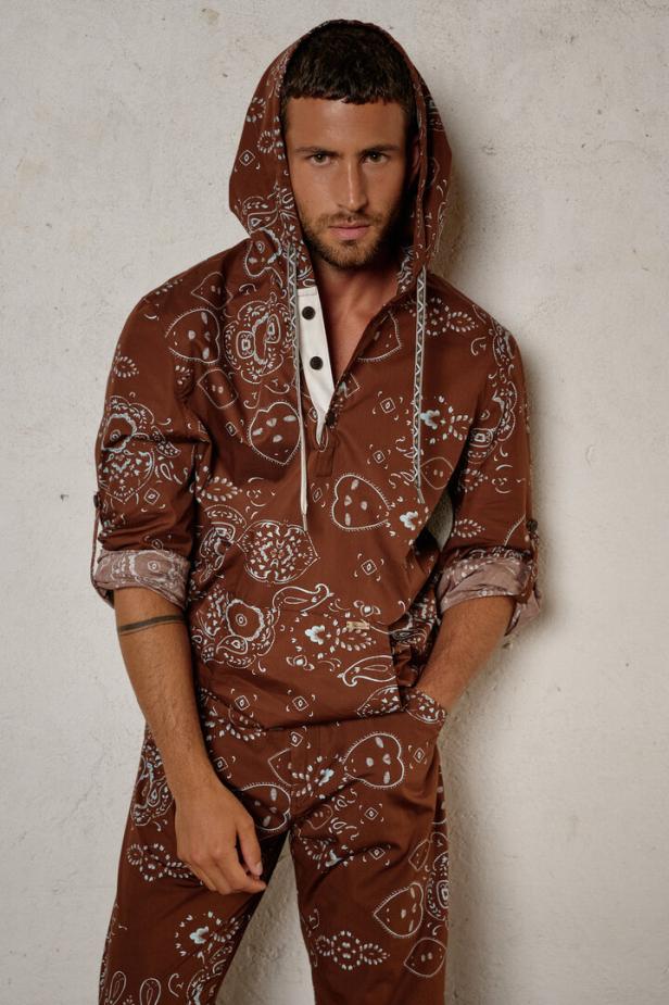 Ein Mann mit Kapuzenpullover und Hose mit braunem Paisley-Muster steht vor einer Wand.