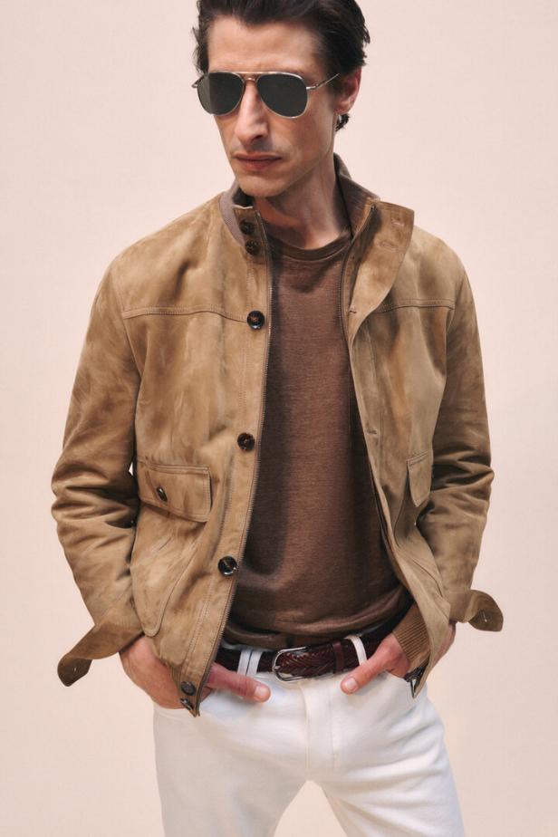 Ein Mann mit Sonnenbrille trägt eine Wildlederjacke, ein braunes T-Shirt und weiße Hosen.