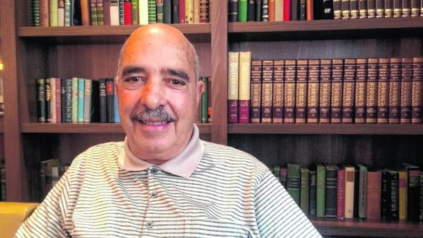 Ein lächelnder Mann mit Schnurrbart sitzt vor einem Bücherregal.