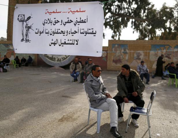 Zwei Männer sitzen auf Stühlen vor einem Banner mit arabischer Schrift.