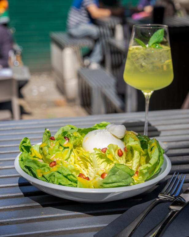 Ein Teller Salat mit Burrata und Granatapfelkernen steht auf einem Tisch, dazu ein Getränk.
