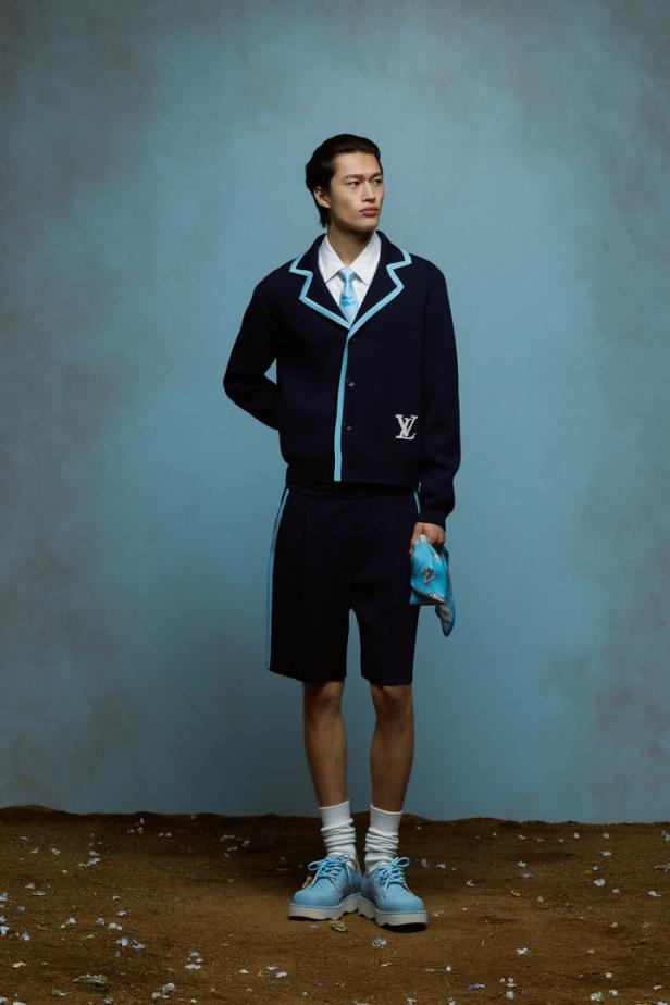 Ein Mann in marineblauer Louis Vuitton-Jacke und Shorts mit hellblauen Akzenten vor blauem Hintergrund.