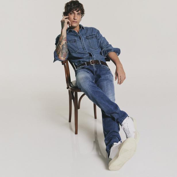 Ein Mann mit Tätowierungen sitzt auf einem Stuhl, gekleidet in Jeanshemd und Jeans.