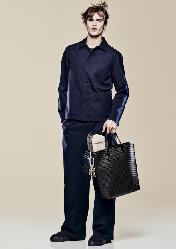 Ein Mann in marineblauem Anzug hält eine schwarze Tasche mit Schlüsseln und Zeitungen.