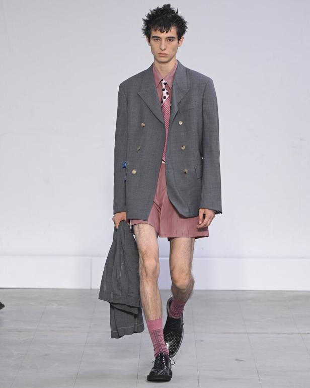 Ein Mann auf dem Laufsteg trägt einen grauen Blazer und Shorts mit rosa Hemd und Krawatte.
