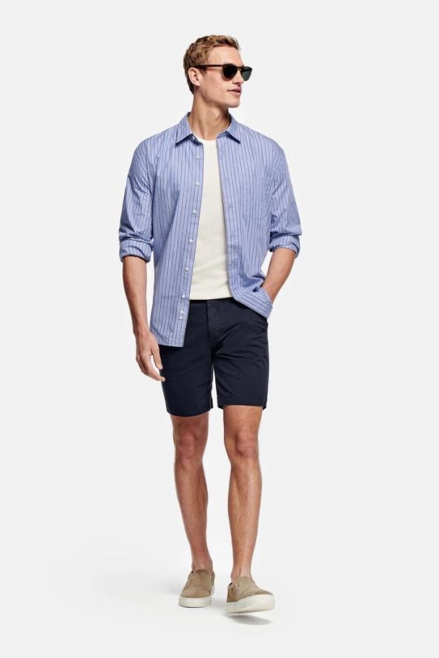 Ein Mann mit Sonnenbrille, blau gestreiftem Hemd und Shorts steht vor einem weißen Hintergrund.