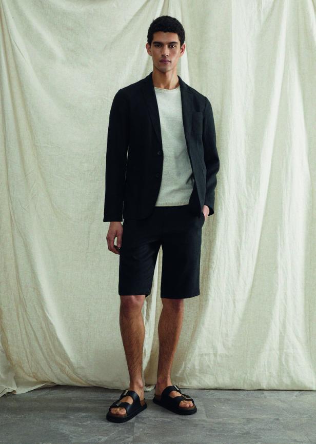 Ein Mann trägt einen schwarzen Blazer, Shorts und Sandalen vor einem beigen Vorhang.