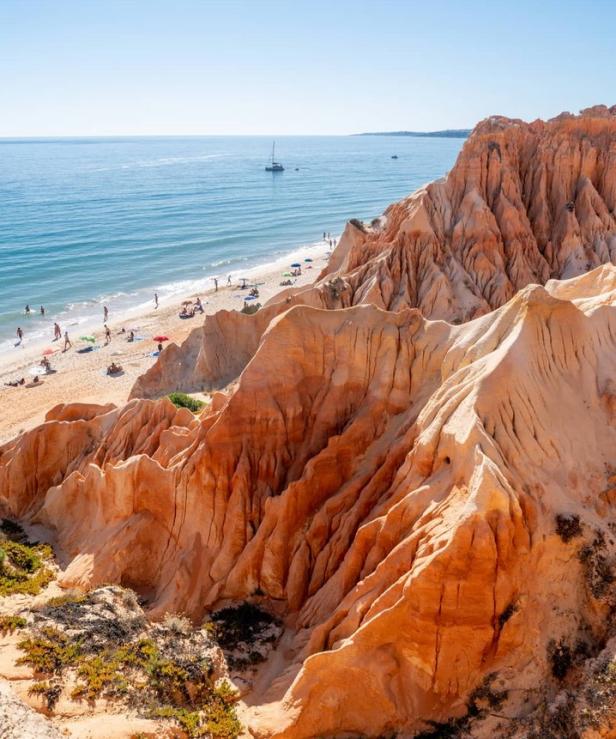 Blick auf einen Strand mit Badegästen und einer markanten, orangefarbenen Felsformation.