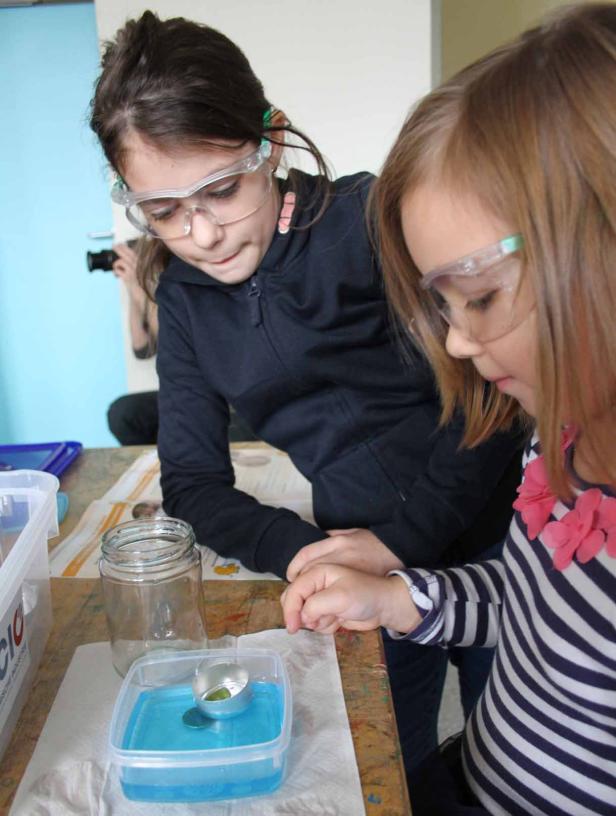 Zwei Mädchen mit Schutzbrillen betrachten ein Experiment mit farbiger Flüssigkeit.