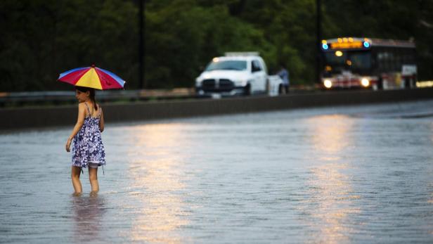 Ein Mädchen steht mit einem bunten Regenschirm im knietiefen Wasser einer überfluteten Straße.