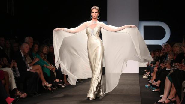 Eine Frau präsentiert ein langes, silbernes Kleid mit einem weiten, transparenten Überwurf auf einem Laufsteg.