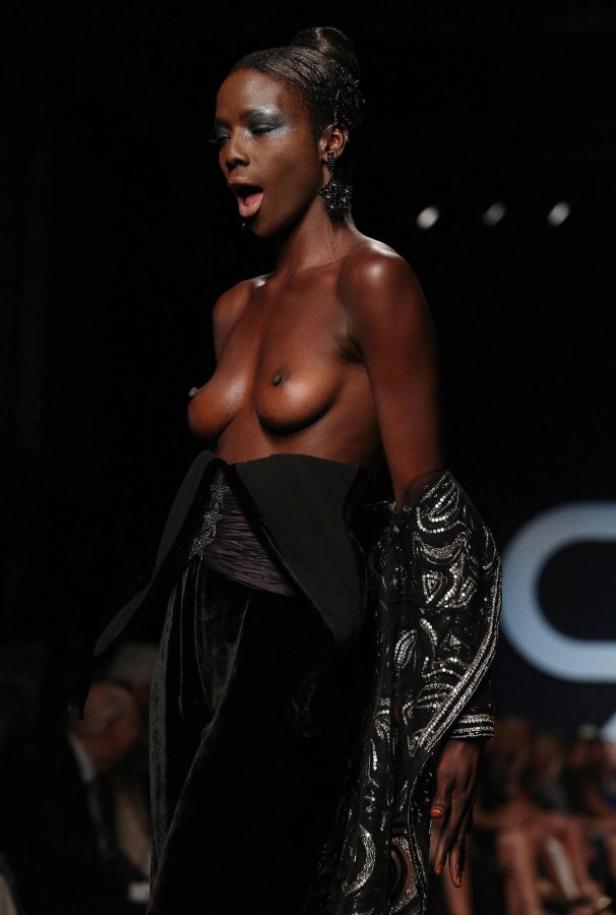 Eine dunkelhäutige Frau mit offenem Mund auf einem Laufsteg, bekleidet mit einem schwarzen Outfit.