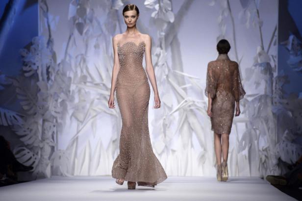 Eine Frau präsentiert ein transparentes, mit Perlen besetztes Abendkleid auf dem Laufsteg.