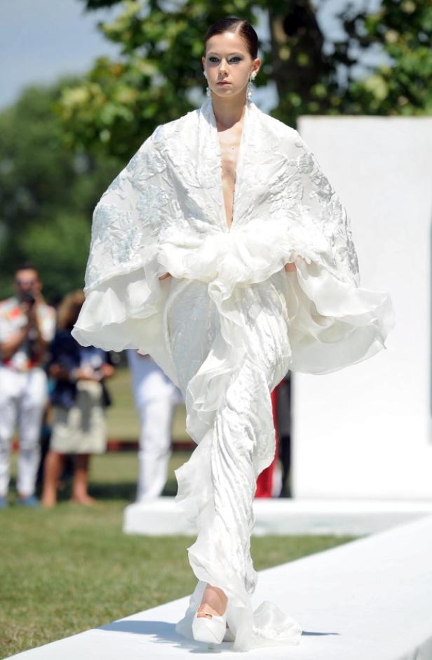 Eine Frau präsentiert ein weißes, fließendes Kleid auf einem Laufsteg im Freien.