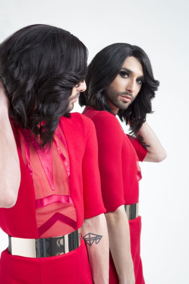 Conchita Wurst posiert in einem roten Kleid mit goldenem Gürtel vor einem Spiegel.