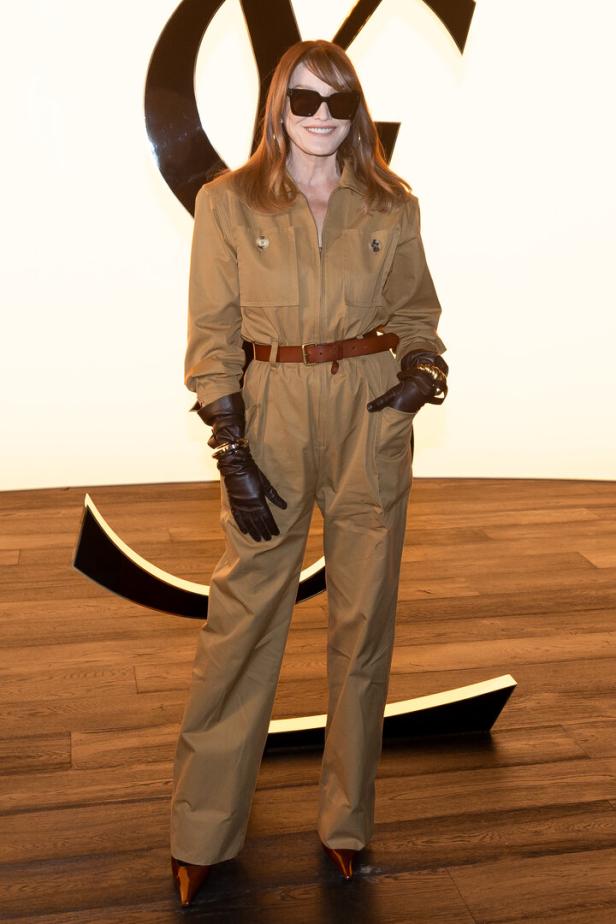 Carla Bruni trägt einen Overall, braune Handschuhe und eine Sonnenbrille vor einem YSL-Logo.