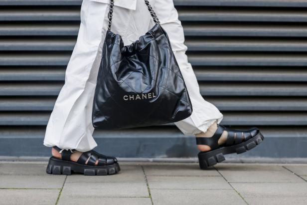 Eine Person mit weißen Hosen und schwarzen Sandalen trägt eine schwarze Chanel-Tasche.