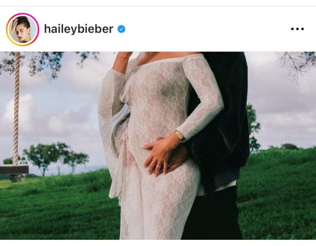 Hailey Bieber in einem weißen Kleid, umarmt von einem Mann vor einer grünen Landschaft.