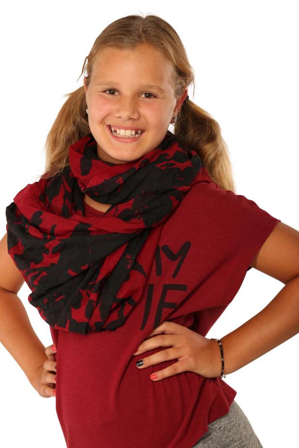 Ein junges Mädchen mit Zöpfen und einem roten T-Shirt mit einem schwarzen und roten Schal lächelt.