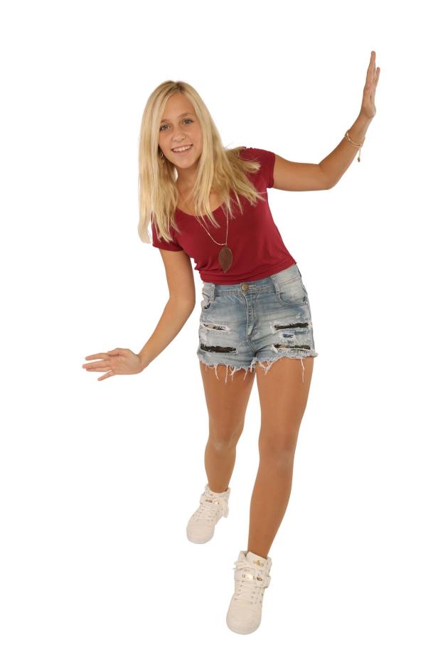 Eine junge Frau in Jeansshorts und einem roten T-Shirt posiert lächelnd vor einem weißen Hintergrund.