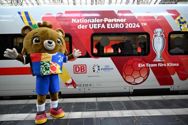Eine Person trägt ein Maskottchen-Kostüm, das einem Teddybären ähnelt. Die Figur steht vor einem Zug der Deutschen Bahn, auf dem Fußball und Pokal aufgemalt sind.