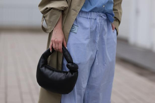 Eine Frau hält eine schwarze Prada-Handtasche; sie trägt einen Trenchcoat und blau-weiß gestreifte Hose.