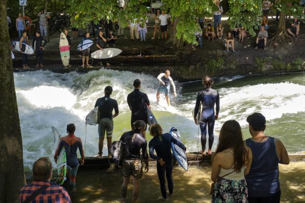 Surfer reitet auf einer Welle in einem Fluss, während andere Surfer und Zuschauer am Ufer stehen.
