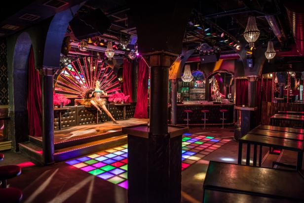 Eine Tänzerin sitzt auf einer Bühne in einem Nachtclub mit farbiger Beleuchtung.