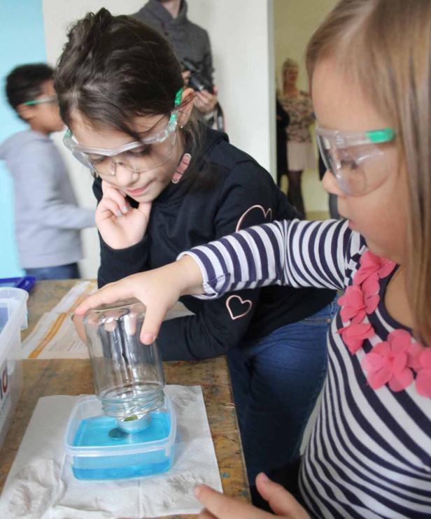 Zwei Mädchen mit Schutzbrillen führen ein Experiment mit einem Glas und einer blauen Flüssigkeit durch.