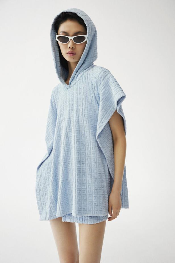 Eine Frau mit Sonnenbrille trägt einen hellblauen, gemusterten Kapuzenponcho und Shorts.