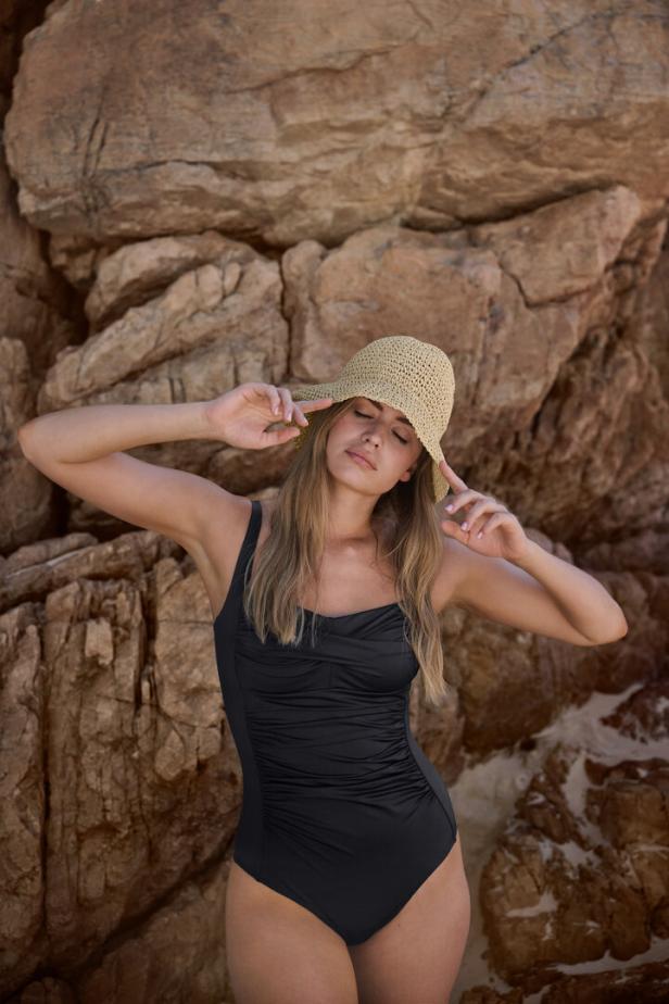 Eine Frau mit Hut und schwarzem Badeanzug posiert vor einem Felsen.
