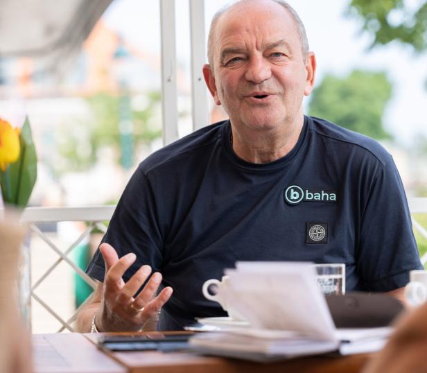 Ein Mann mit einem T-Shirt der Marke „baha“ sitzt an einem Tisch im Freien und gestikuliert.