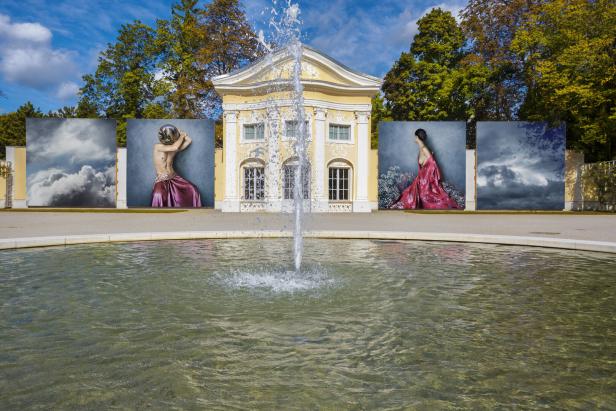 Ein Springbrunnen vor einem gelben Gebäude, flankiert von großformatigen Kunstfotografien.