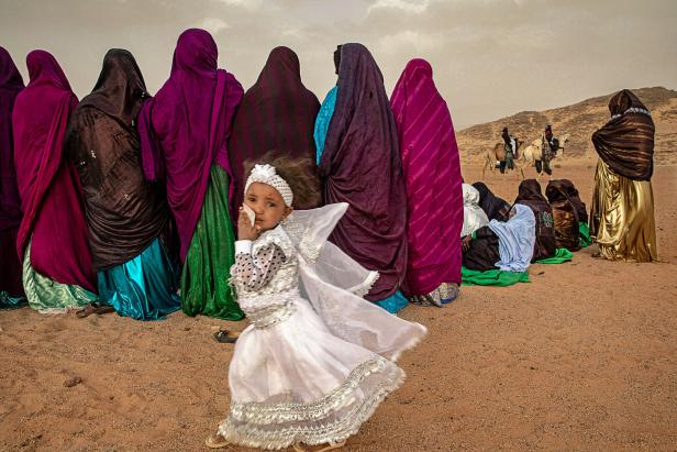 Ein junges Mädchen in festlicher Kleidung steht vor einer Gruppe verschleierter Frauen in einer Wüstenlandschaft.