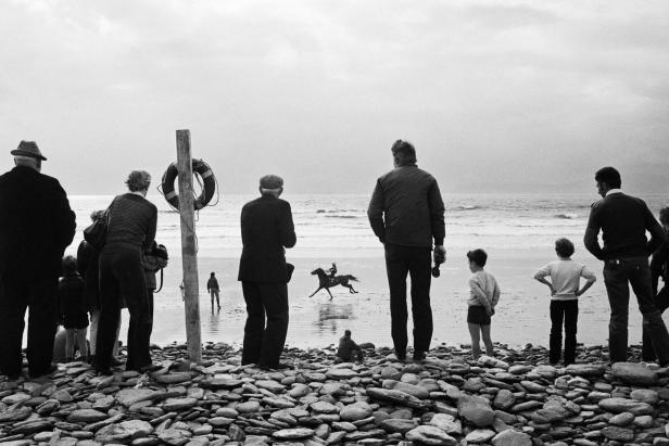 Eine Gruppe von Menschen beobachtet ein Pferderennen am Strand.