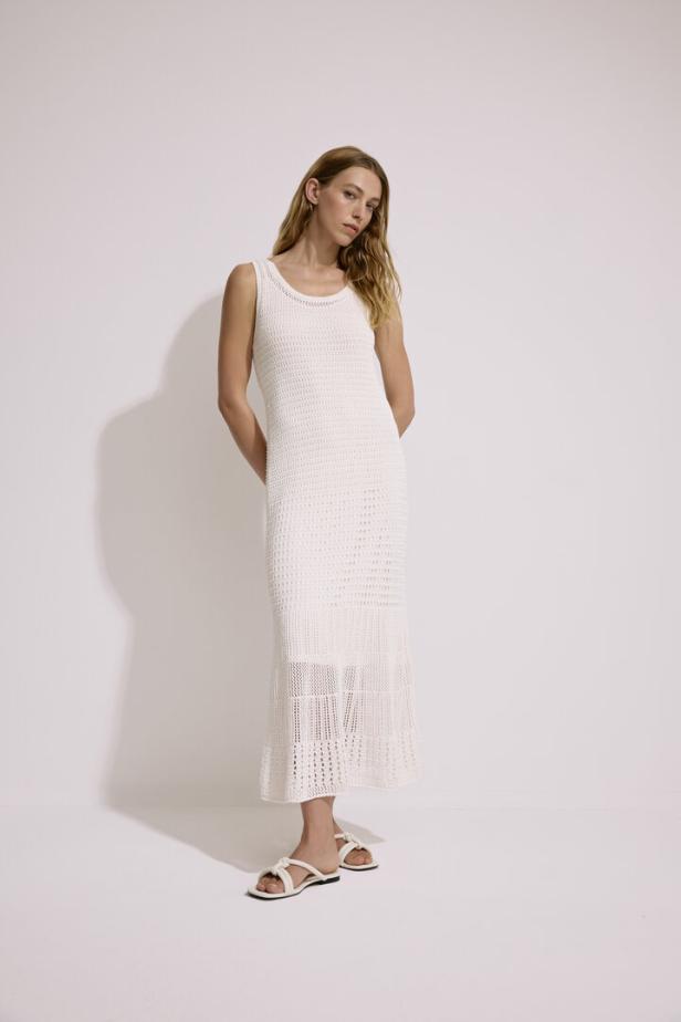 Eine Frau trägt ein ärmelloses, gehäkeltes, weißes Maxikleid und Sandalen.
